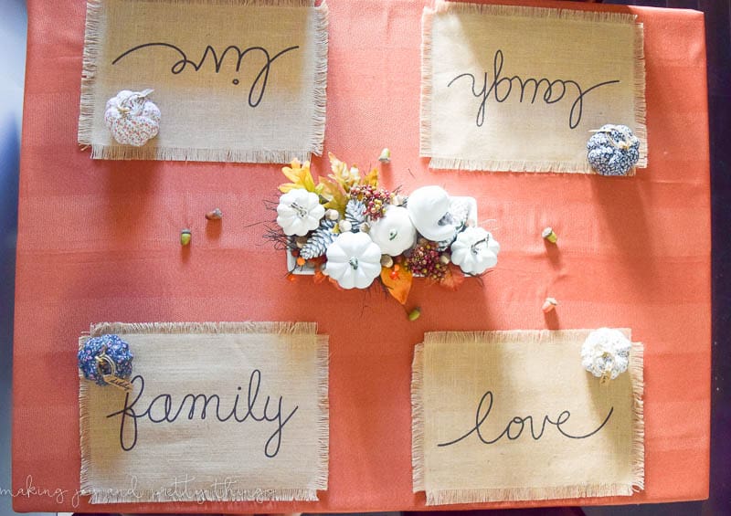 An overhead look at the fall tablescape in our dining room. The table is dressed with a burnt orange table cloth and a fall centerpiece made with faux white pumpkins, acorns, pinecones, and leaves. Each seat has a burlap placemat and a patterned faux pumpkin. The placemats each say "family", "love", "laugh", and "live"