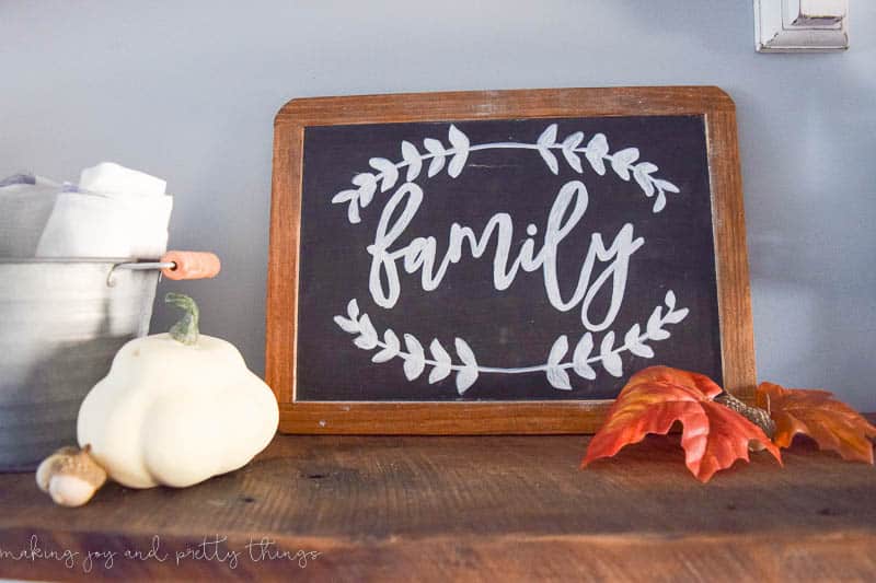 A wood-framed chalkboard sign that says "family" in white chalk sits on a wood shelf, surrounded by faux orange leaves and a faux squash.