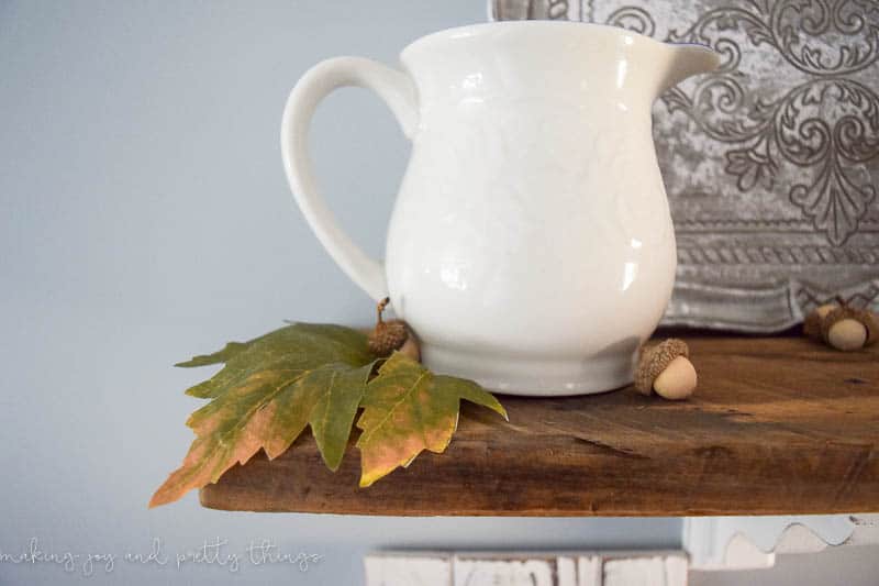 A white milk jug vase sits on a wooden shelf surrounded by faux leaves and mini acorns. 