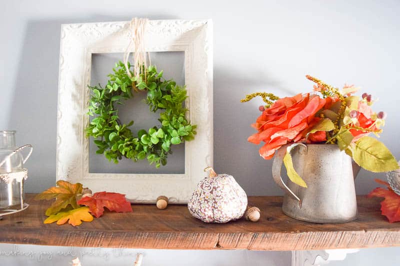 A dining room shelf contains assorted fall decor. A metal can holds faux flowers and leaves. There is a white picture frame with a faux greenery wreath hanging from twine in the center. Faux leaves and mini acorns also sit on the shelves.