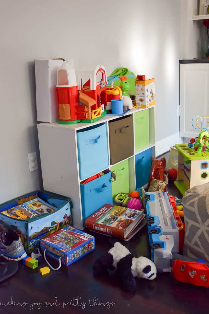 A messy corner of the living room, cluttered with kids toys and books. A six-cube shelf with blue, green and brown cloth baskets holds more kid toys.
