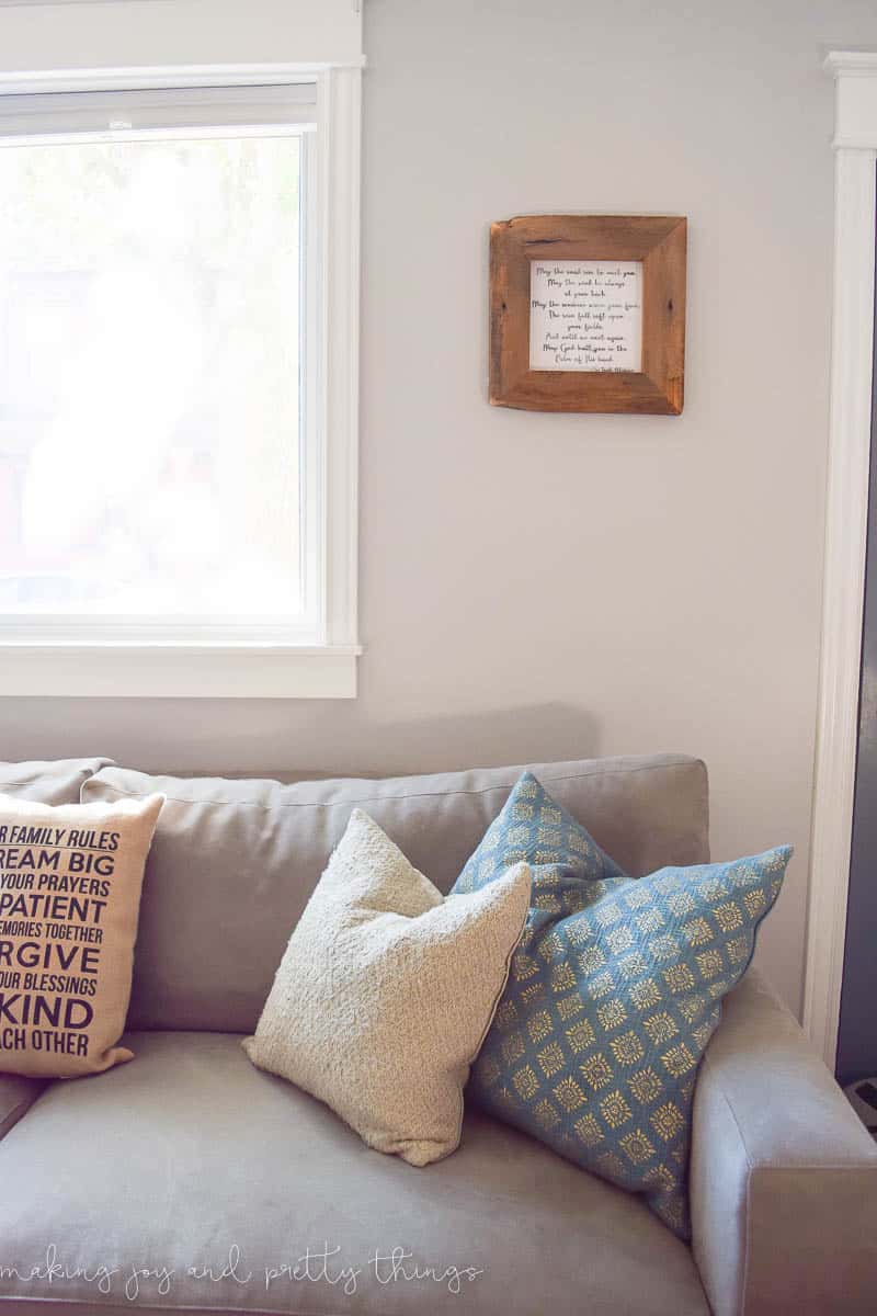 A living room with a gray suede couch decorated with blue and tan throw pillows. Above the couch hangs a rustic wood picture frame and print of a poem.