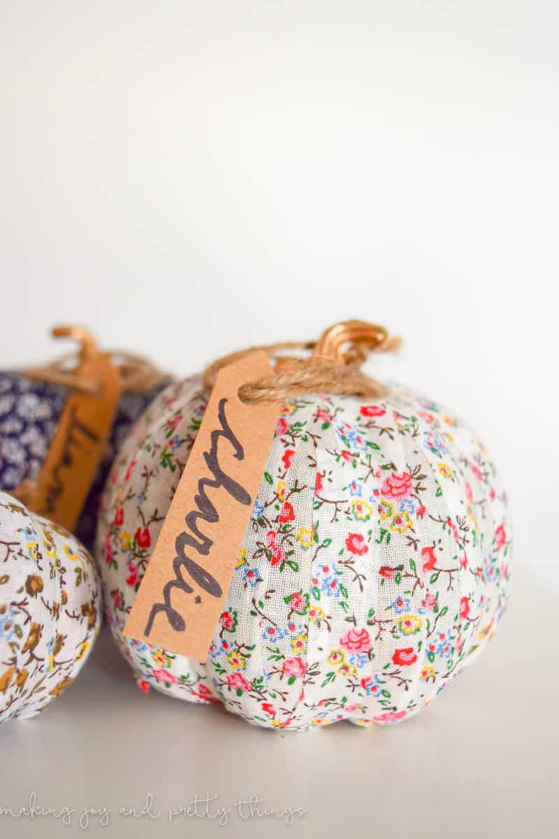 A small fabric pumpkin with a colorful floral pattern has a handwritten name tag attached to the stem.