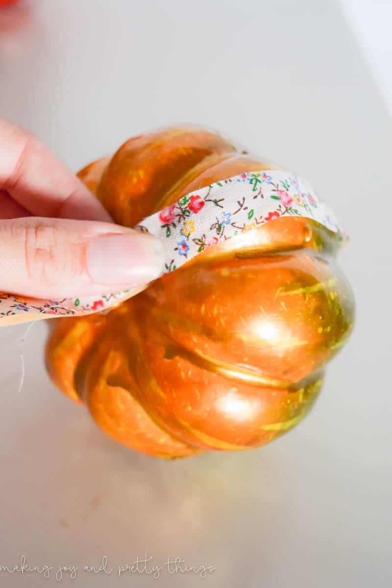 A woman's hand takes a strip of floral washi tape and attaches it around a small orange craft pumpkin.