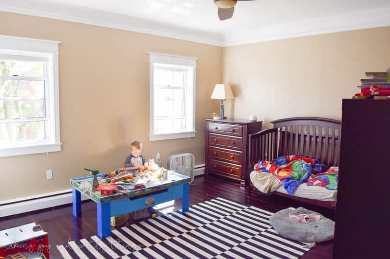 A corner of a boys bedroom with tan walls, a dark wood dresser, a play area with a toy table, and a black and white striped area rug.