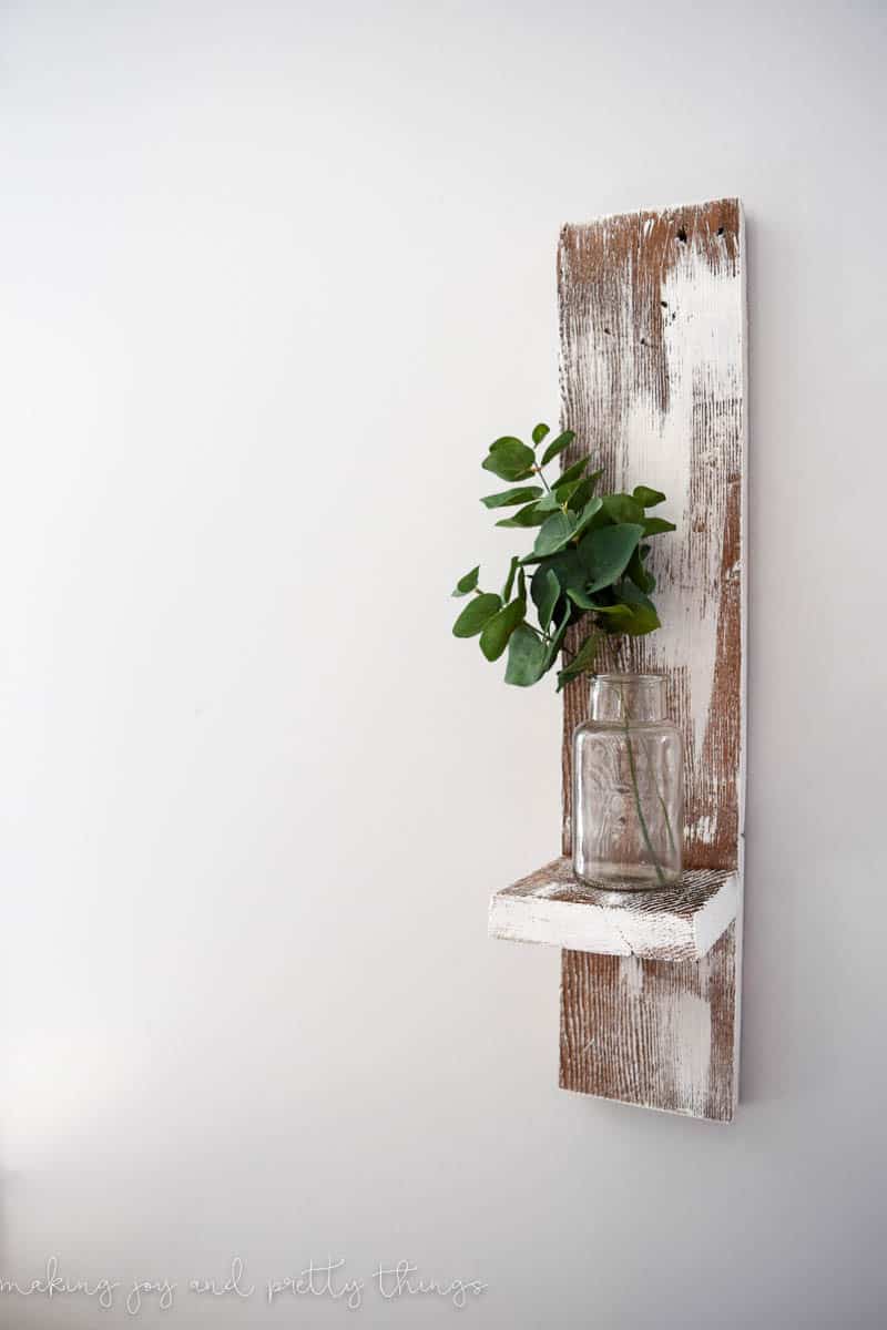 Small vertical shelf for wall made with barn wood holding a small clear vase 
