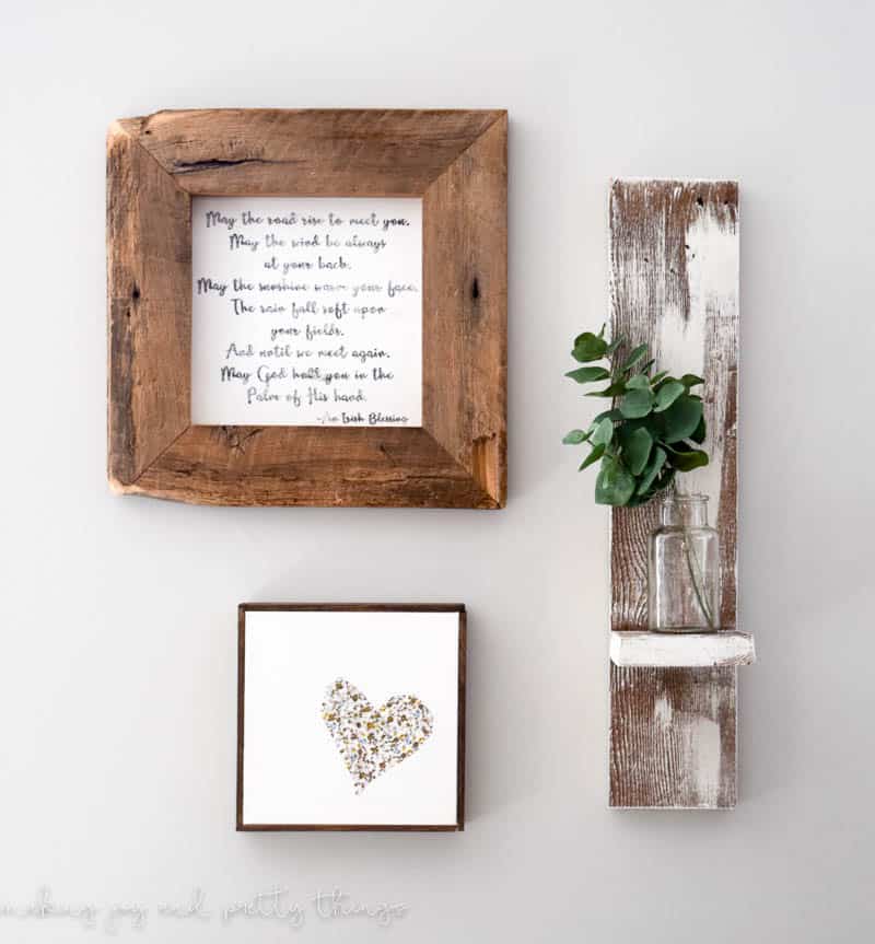 Mini gallery wall with irish blessing sign with a frame made from barn wood, a small white canvas with heart design and dark wood frame along with a reclaimed wood shelf holding a small vase with white wash finish