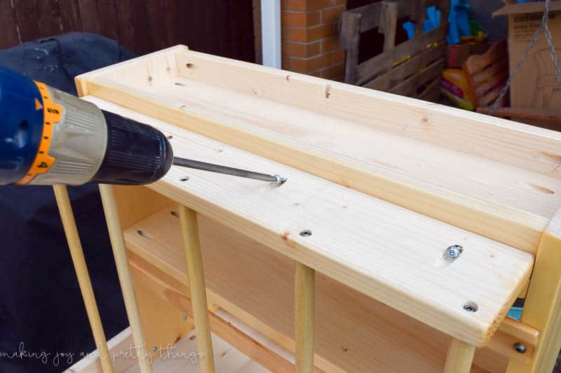 A person uses a drill to insert screws into the pocket screw holes to attach the book holder to the table side shelves.