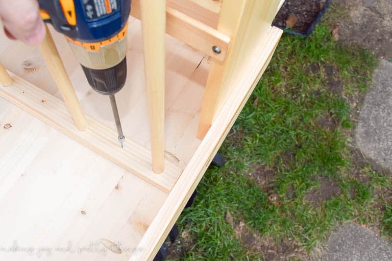 A person uses an electric drill to drill screws into the wooden book holder, attaching it to the underside of the table.
