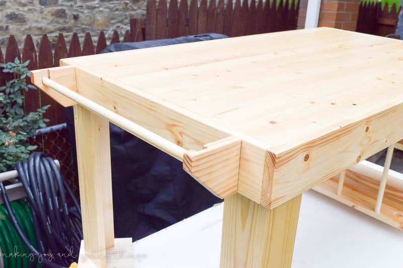 A paper holder attached to the side of the unstained wooden craft table. The paper holder is made from two ends and a long wooden dowel rod to hold reems of paper.