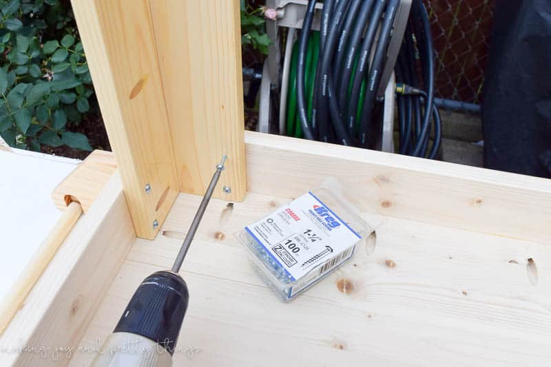 Using a drill to insert screws into the legs of the craft table. A pack of 1-1/4 inch screws sits next to the drill.