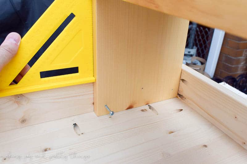 A hand holds a yellow right-angle ruler to make sure the table legs are perfectly square with the rest of the table.