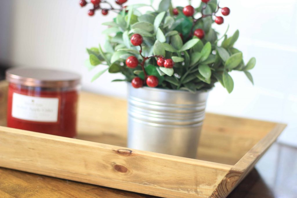 A lightly stained rustic wood tray sits on a darker stained wooden table. A jarred candle and a holly berry bush in a milk can tin sit within the wooden tray.