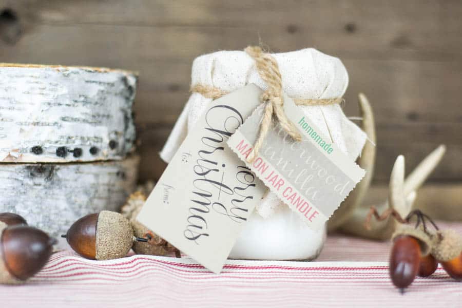 A homemade mason candle jar sits on a table, surrounded by decorative acorns. The mason jar has DIY gift tags that say "merry Christmas" and "homemade vanilla mason jar candle". 