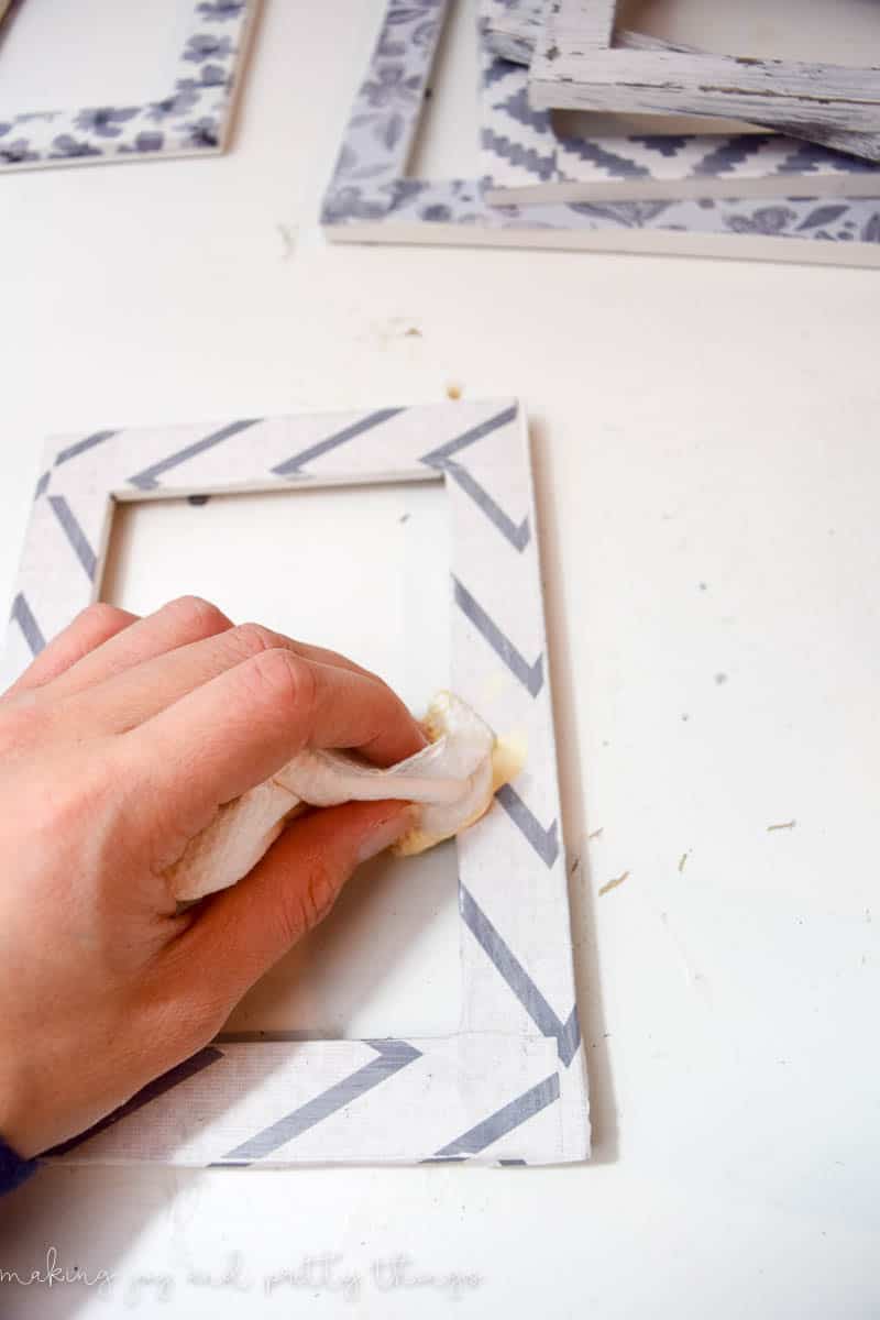 A hand holding a paper towel is wiping distressing wax over the surface of a paper-covered picture frame to give it a distressed farmhouse look. 