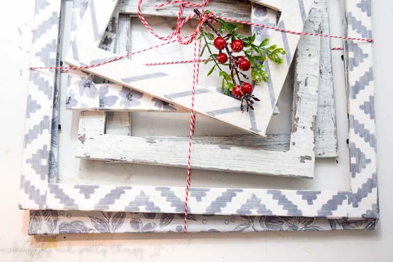 A stack of DIY wooden picture frames sits on a white table, tied together with string and a holly berry bunch. The picture frames are different sizes, and painted white with gray patterns around the frames.