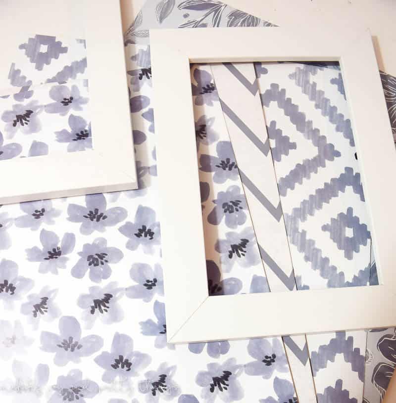 Two white wood picture frames sit on top of sheets of craft paper, each with a different white and gray design.