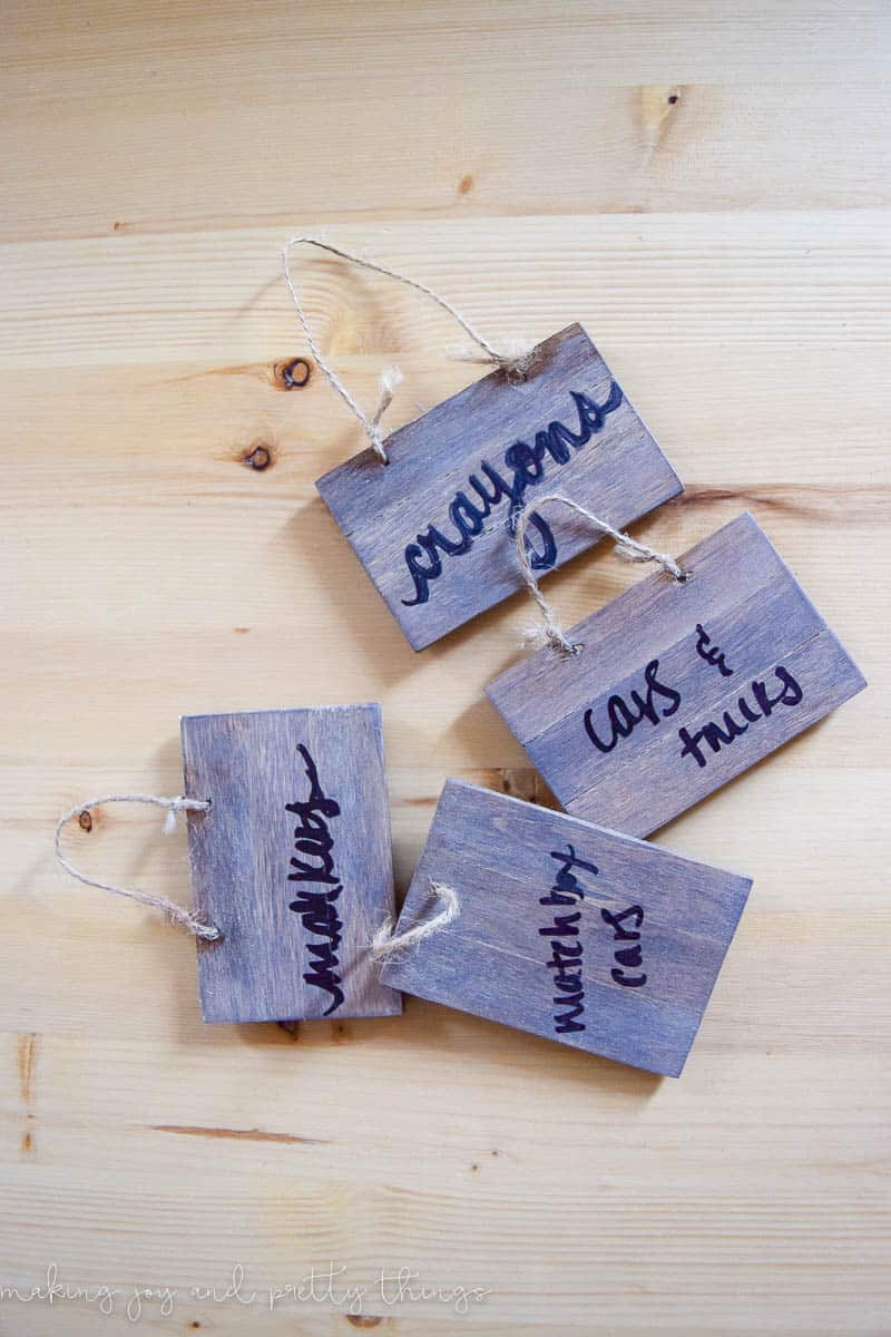 Four small wood DIY basket labels with a whitewashed wood effect sit on a wood table. Each has a different handwritten label - markers, matchbox cars, cars & trucks, and crayons.