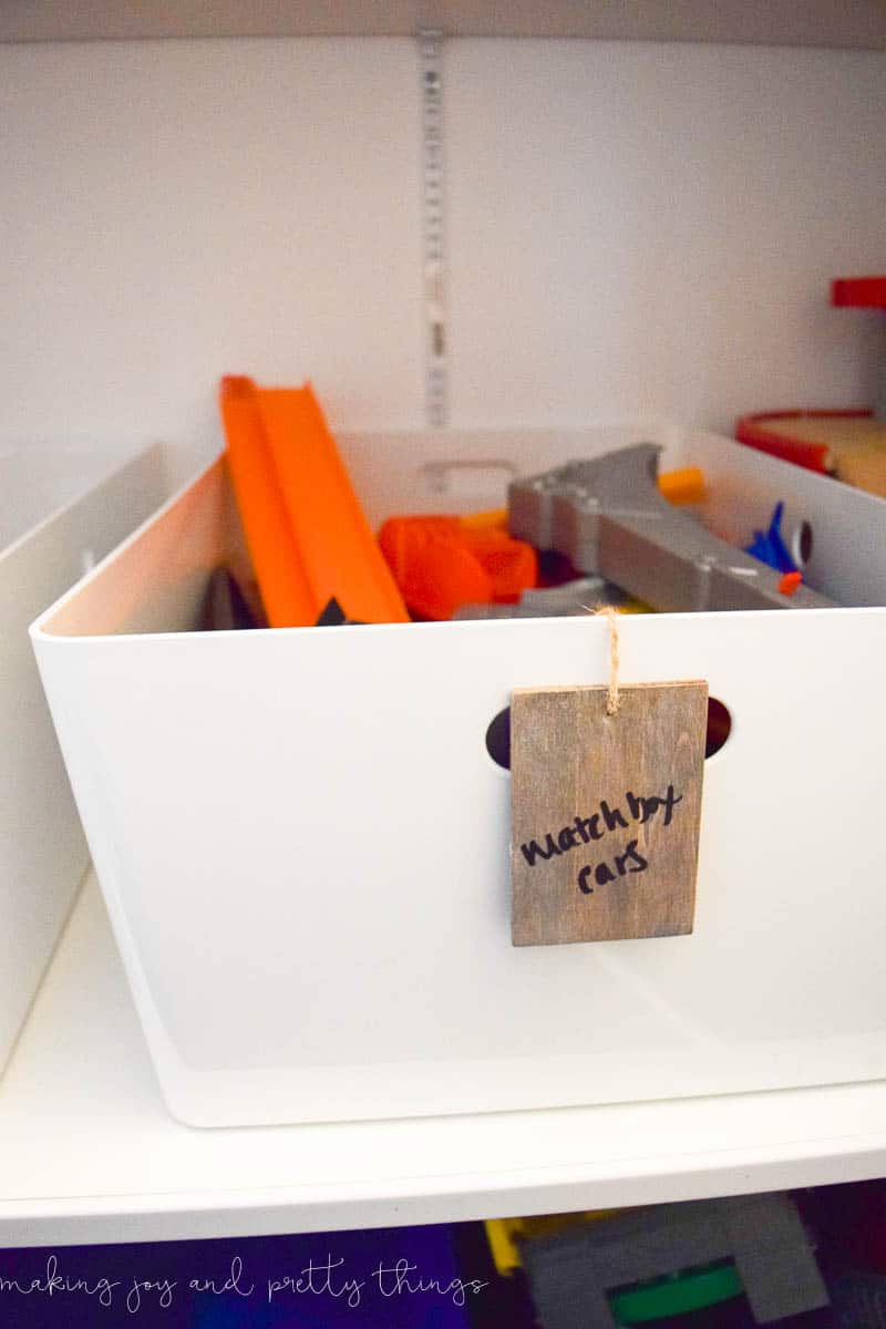 A white storage bin filled with toys sits on a shelf. A small wood DIY basket label hangs on the handle with a piece of twine. The label reads "matchbox cars" in handwritten black marker.