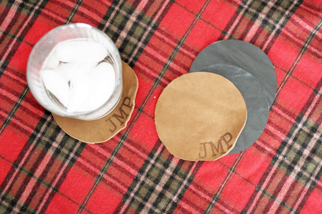 Personalized leather coasters with embossed letters sit on a red, black and white plaid tablecloth. The coasters are made from tan and dark gray colored leather. A glass with liquid and ice sits on one coaster.
