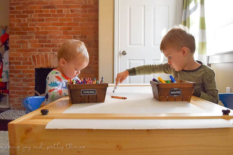 Toy young boys sit at a small wooden crafting table, coloring on paper with bins of crayons and markers.