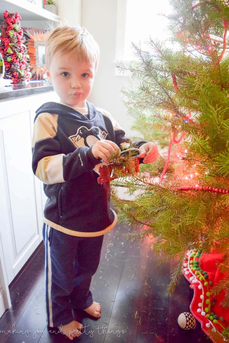 A young boy with a sassy smirk on his face hangs a Christmas ornament onto a branch of a Christmas tree.