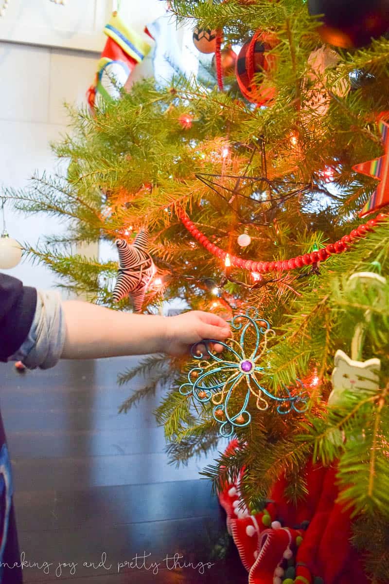 A close up look at a decorated Christmas tree, adorned with sparkling Christmas lights, garland, and ornaments. A young boy's arm reaches out and holds a unique star ornament hanging at the bottom of the tree.