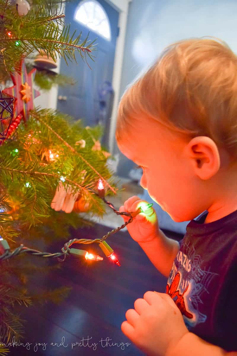 A little boy plays with a string of red and green Christmas lights, attached to the bottom branches of a Christmas tree. His face is bathed in red light from the tree lights.