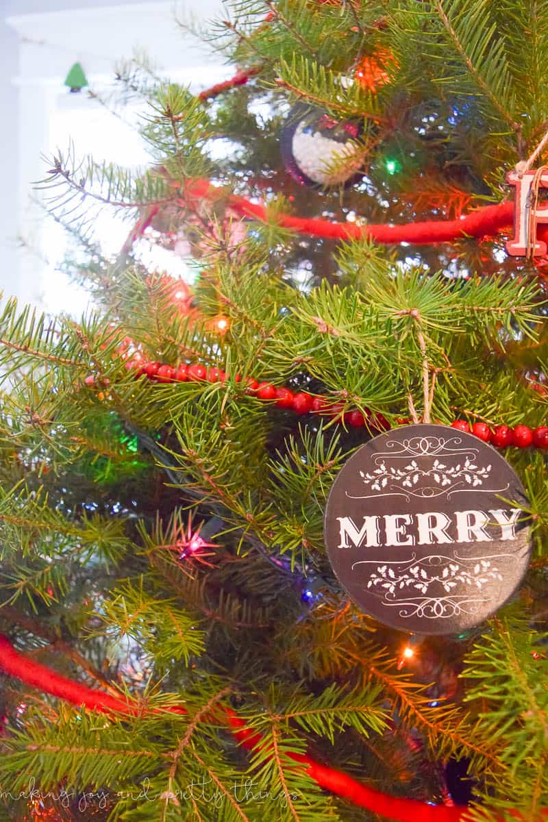 A close up image of the branches of a Christmas tree, decorated with sparkling lights and red bead garland. A round ornament hangs from a branch and says "MERRY"
