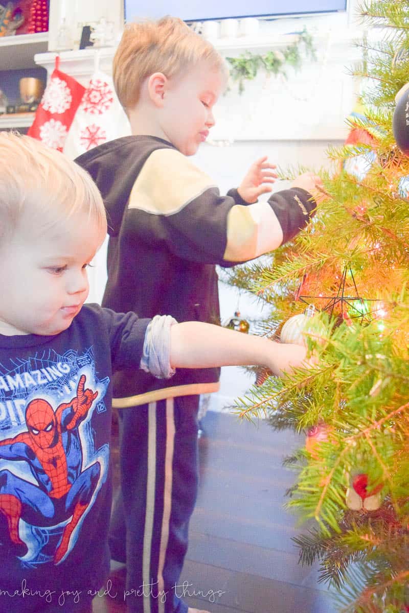 Two young toddler boys helping to decorate a Christmas tree by hanging Christmas ornaments.