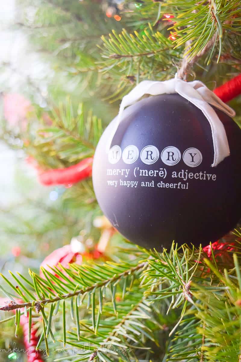 A black round ornament hangs from the Christmas tree branches. It's printed with the word MERRY, with the definition below; it says "merry - adjective: very happy and cheerful"