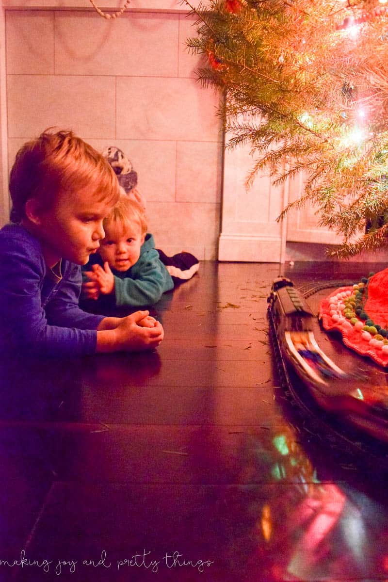 Two young boys sit on the floor, watching a toy train run on train tracks around the Christmas tree. The soft light from the Christmas tree lights cast a red glow in the space.