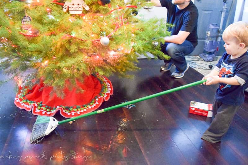 A young boy holds a broom, helping to clean up fallen pine needles from a decorated Christmas tree.
