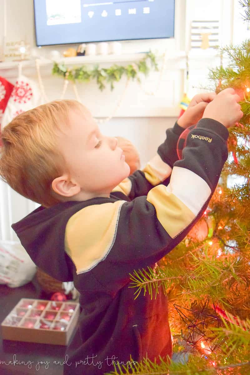 A young boy carefully hangs Christmas ornaments onto the branches of a Christmas tree, sparkling with red Christmas lights.