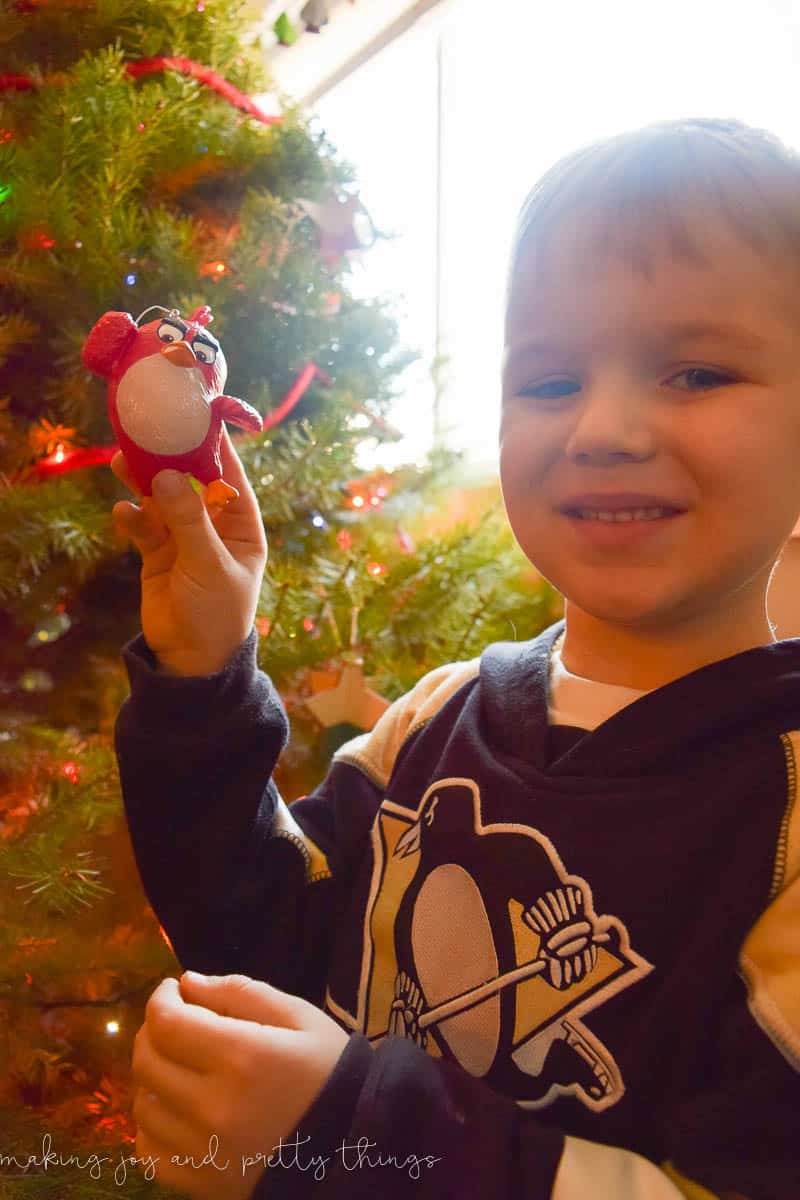 A little boy stands in front of a decorated Christmas tree, proudly holding up an Angry Birds Christmas ornament.