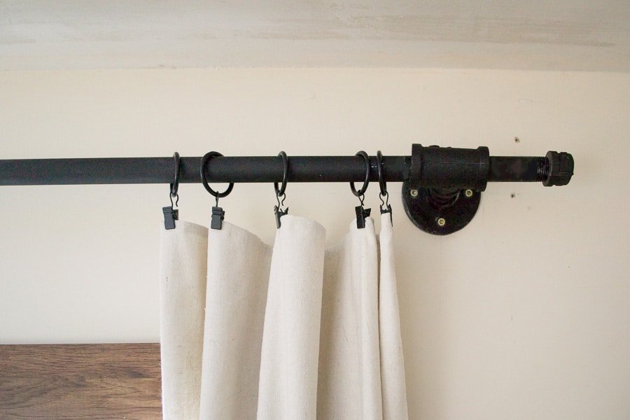 A white linen curtain hangs from curtain clips, strung on to a matte black curtain rod, held up by a wall-mounted metal flange.