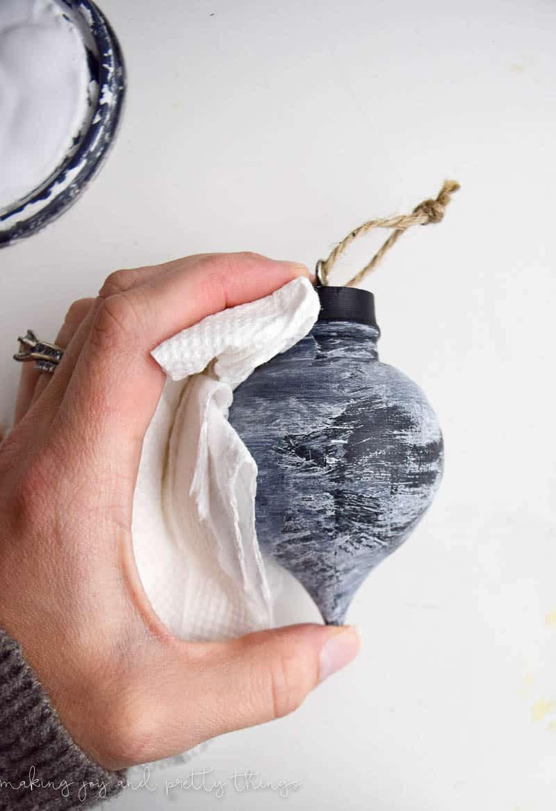 A woman's hand uses a paper towel to wipe white chalk paint off the surface of a black wood ornament, leaving a streaky white paint finish on the ornament,