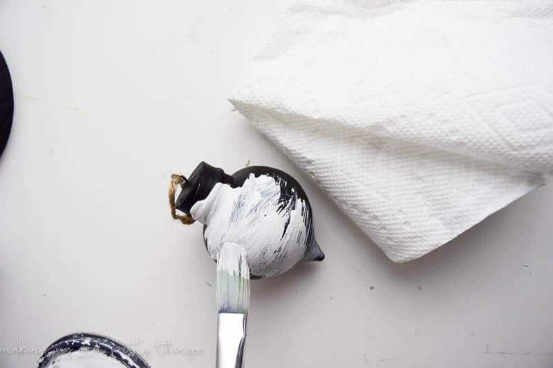 A small black ornament sits on a white tabletop next to a white paper towel. A paint brush applies white chalk paint to the surface of the ornament.