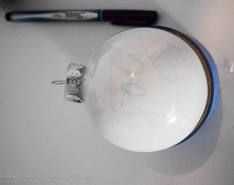 A clear plastic globe ornament is filled with a layer of white chalk paint which coats the inside of the ornament ball. On the outside, the word "joy" is faintly traced onto the surface of the ornament.