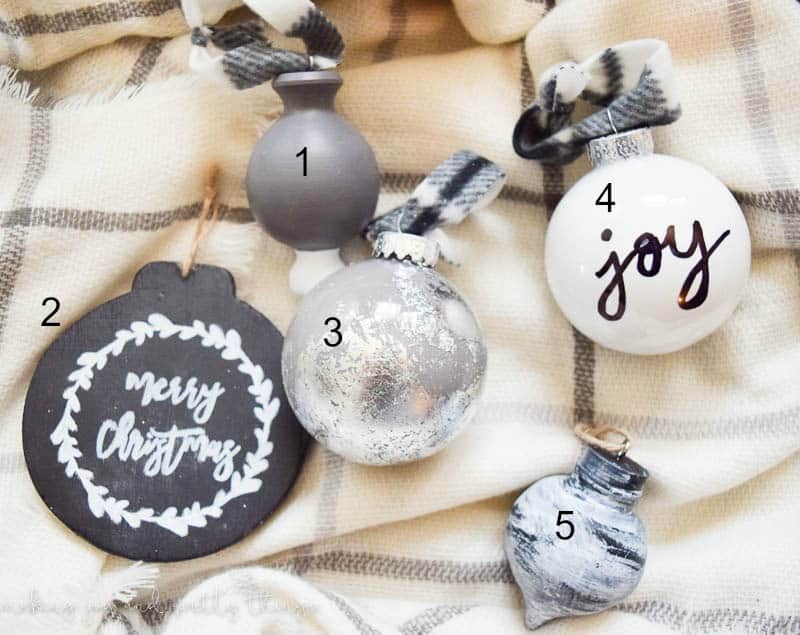 Five DIY painted Christmas ornaments sit on a white and gray plaid blanket. Three of the ornaments are blubs - one is gray, one silver, and one is white with the word "joy" handwritten in black marker. A fourth ornament is a round black disk with "merry christmas" and a laurel wreath painted in white paint. The fifth ornament is a smaller bulb painted with white, black and gray paint in a marbled effect.