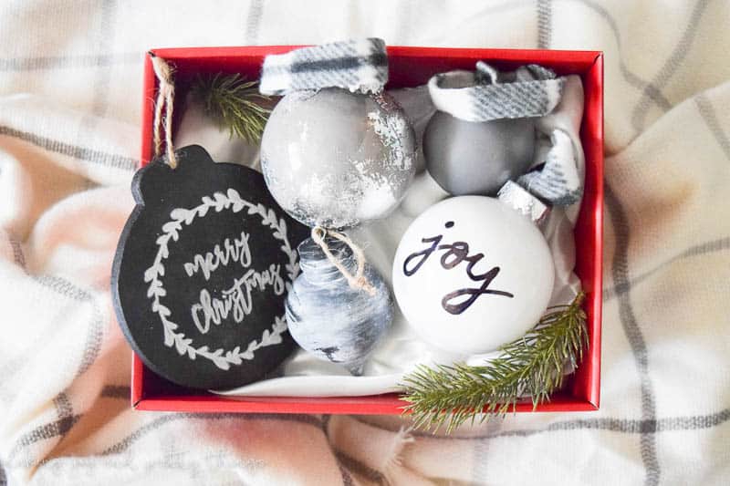 A red gift box sits on a white and gray plaid blanket; the box is filled with DIY hand-painted Christmas ornaments, each decorated with a different design, in the colors black, white, silver, and gray.