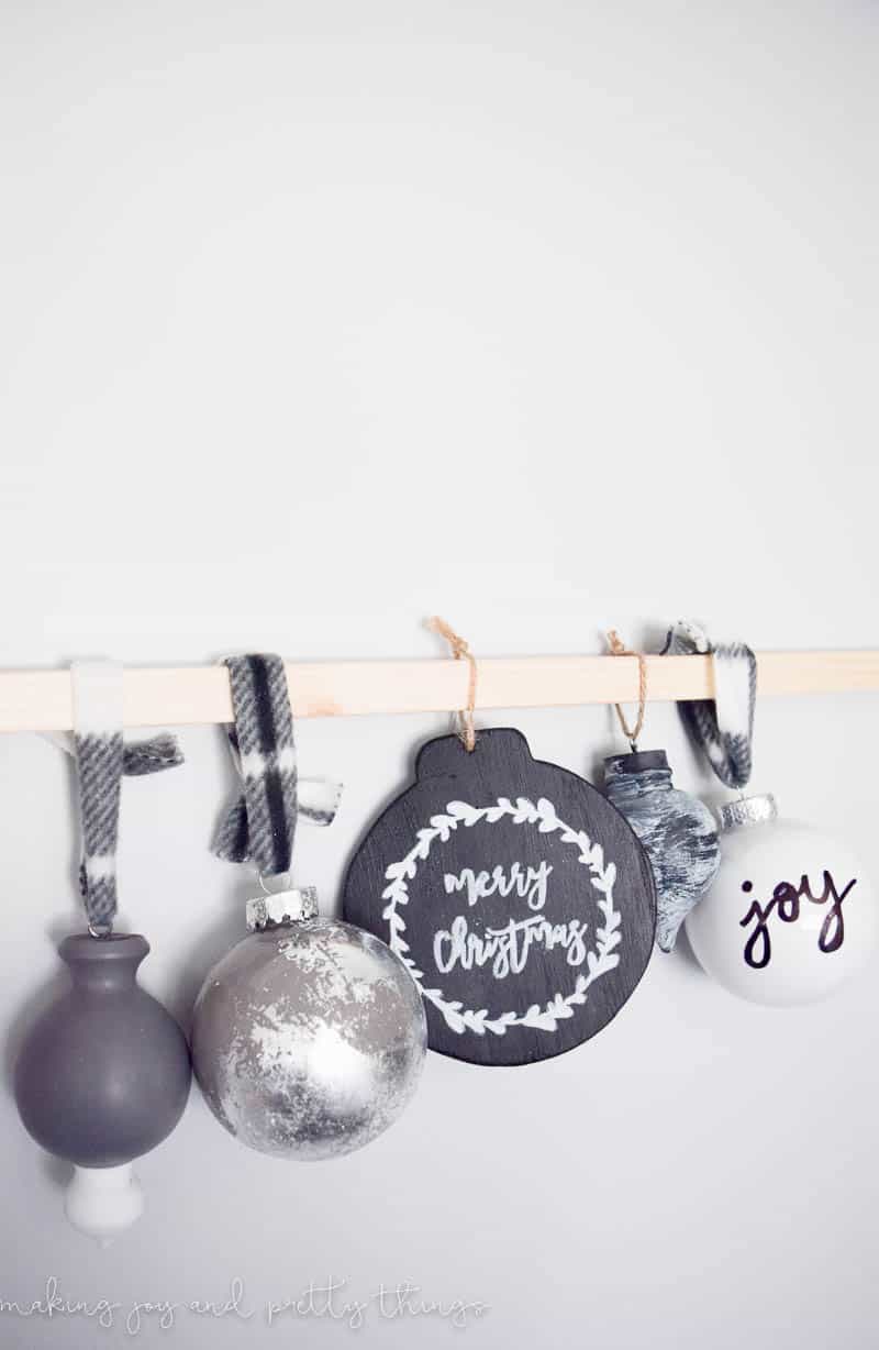 Five DIY painted Christmas ornaments hang on a wooden dowel rod. Three ornaments hang from black and white buffalo check ribbon, and two hang from craft twine. From left to right: a grey and white color-blocked wood ornament; a plastic globe ornament with gray and silver-leaf, a round black chalkboard ornament with a simple wreath and the words "merry christmas" in white; a black wood ornament with a whitewashed paint finish; and a white globe ornament with the word "joy" written in black marker.