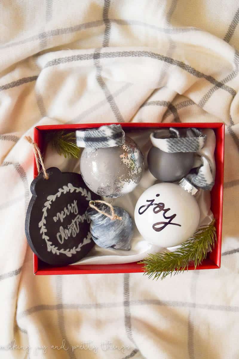 Five homemade painted Christmas ornaments sit in a red box. Each ornament is decorated differently. There are three round globe ornaments in white, gray, and silver. One ornament is a black chalkboard ornament that says "merry christmas", and the fifth ornament is a small black wood ornament with a rustic white-washed paint finish.