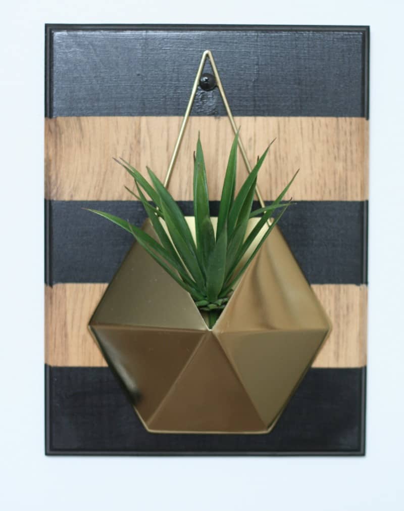 The completed gold and wood wall planter, finished with a faux plant tucked into the hexagon gold planter. The planter is attached to a black and wood-striped board.