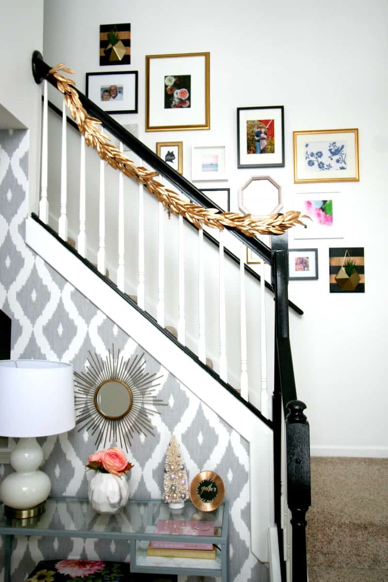A stairway in the entryway of a home, decorated with an eclectic gallery wall. The black stair bannister is decorated with gold leaf garland. The gallery wall is complete with black, white, and gold-framed photos and art in difference sizes. Two gold and wood wall planters hang among the picture frames.