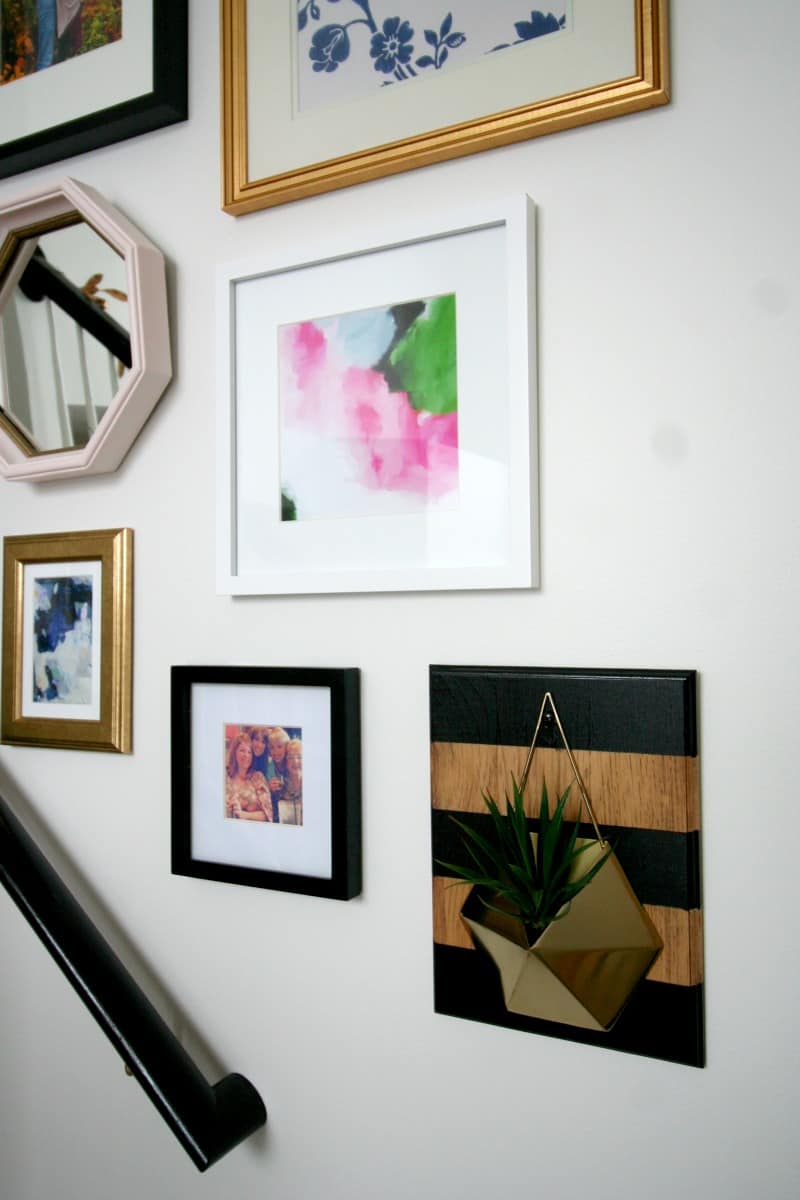 A close up look at one of the glam-farmhouse-style wood wall planters made with a geometric gold planter and black and wood striped board. The planter joins and eclectic gallery wall comprised of black, white, and gold picture frames of various sizes.