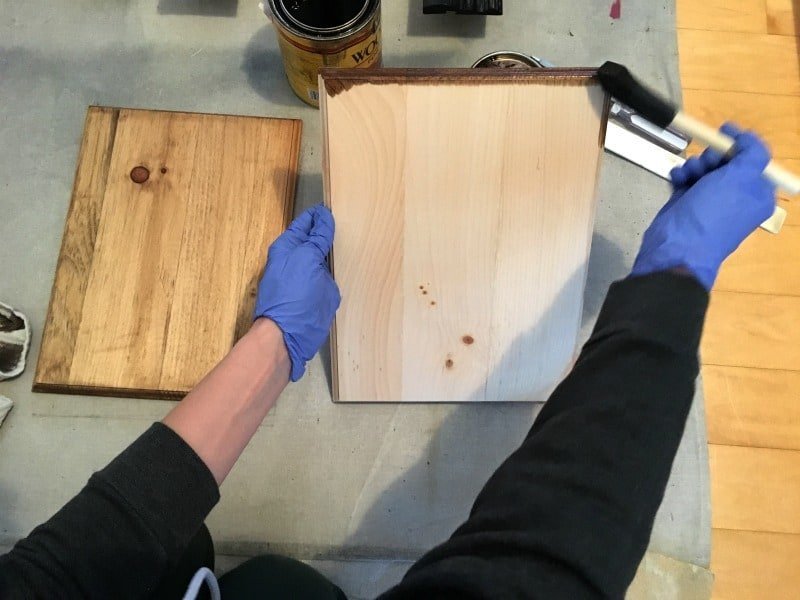 A person applies wood stain to a pine wood board using a sponge brush. A second pine board sits on a drop-cloth covered surface, already stained.