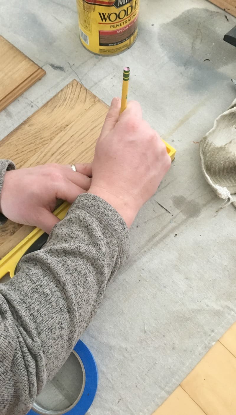 A person uses a measuring tape and pencil to mark out where to put painted stripes on two stained wood boards.