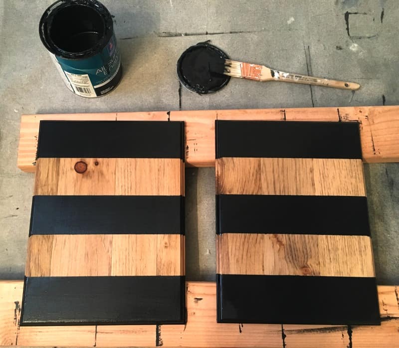 Two pine wood boards are painted with three black stripes and two natural wood stripes per board. And open can of black paint and a paintbrush site beside the wood boards as they dry.