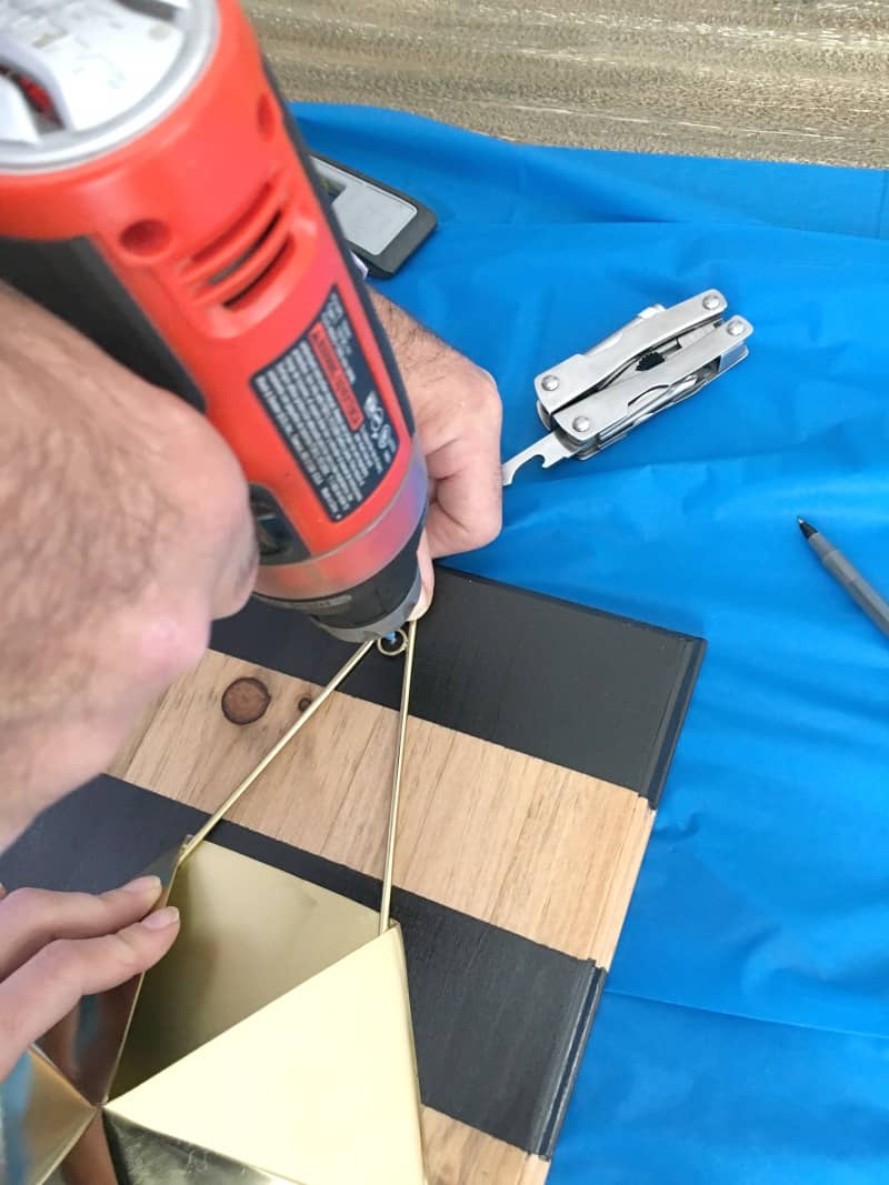 A man uses a power drill to attach the geometric gold planter to a striped wood board.